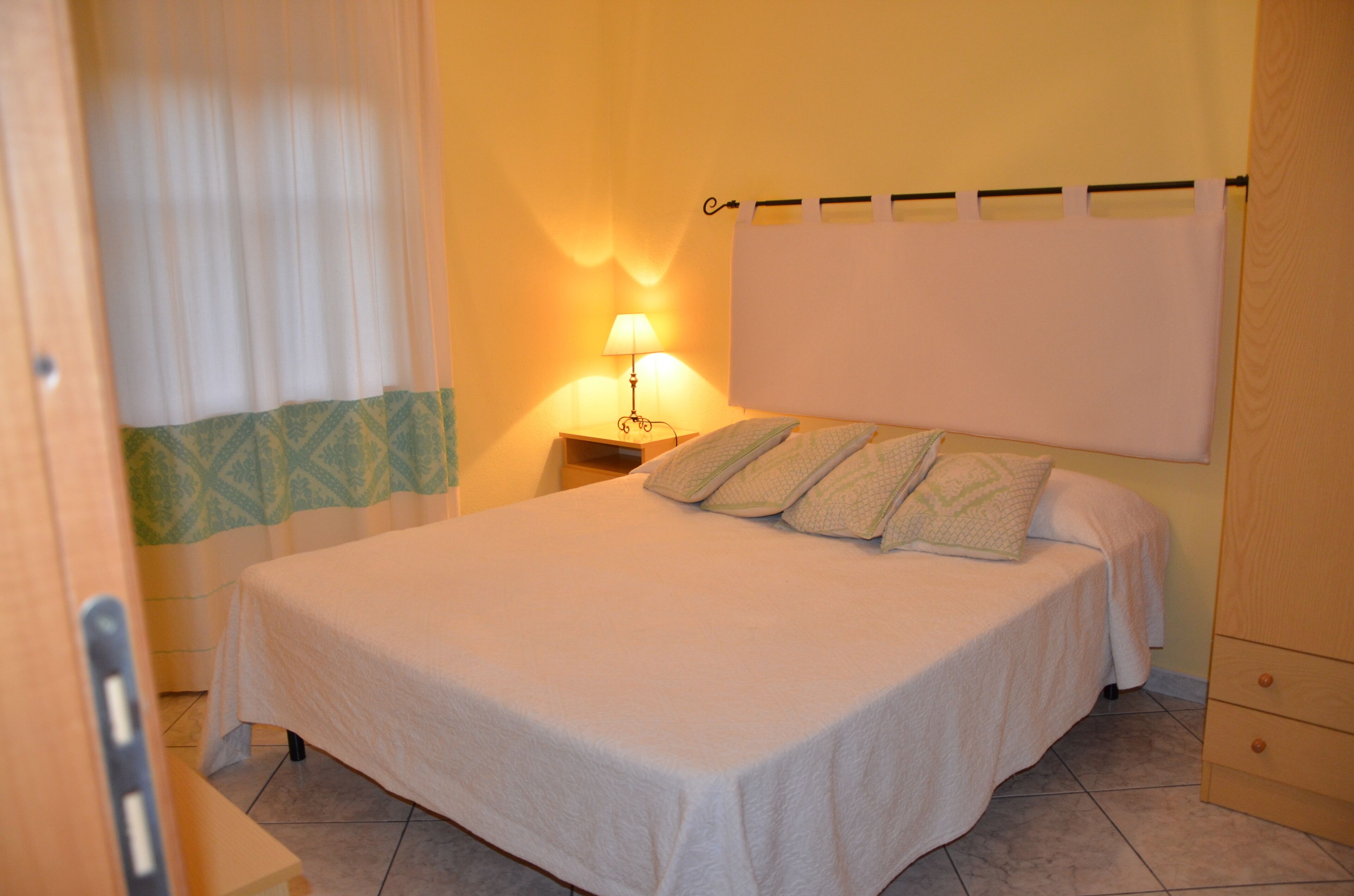 A neatly made bed with a white bedspread and pillows, a bedside lamp, and a curtain partially drawn to the side.