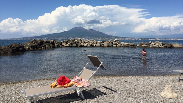 A beach with a rocky breakwater, a lounge chair with a towel and hat, and a person in the water. - For 8 people Monticelli-Mansion (casola di napoli)