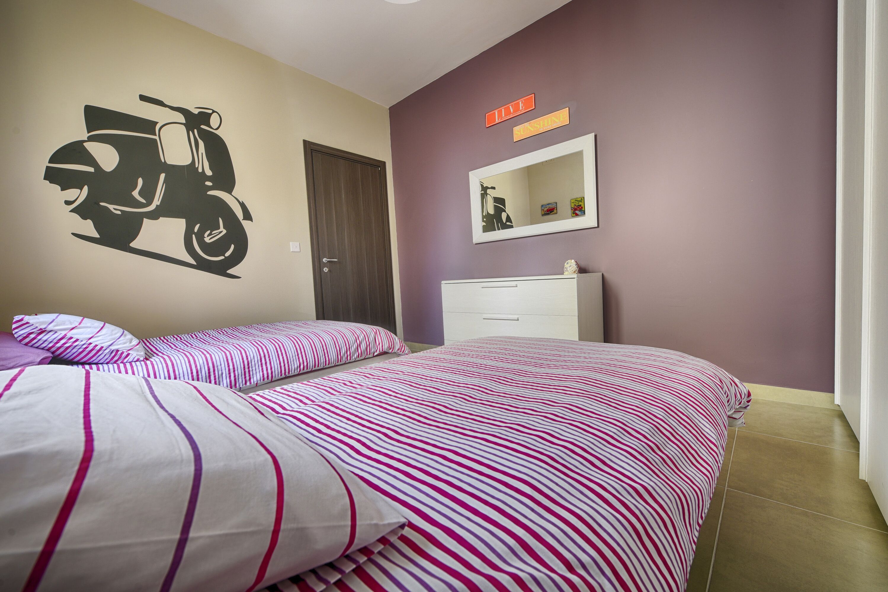 A bedroom with two beds, a wall-mounted mirror, a scooter decal, and a sign that says "LIVE THE EXPERIENCE".