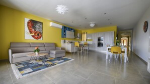 A modern living room with a sofa, coffee table, and rug, adjacent to a kitchen with white cabinets and yellow chairs. - Three bedroom, luxury apartment with private pool (Ix-Xagħra)