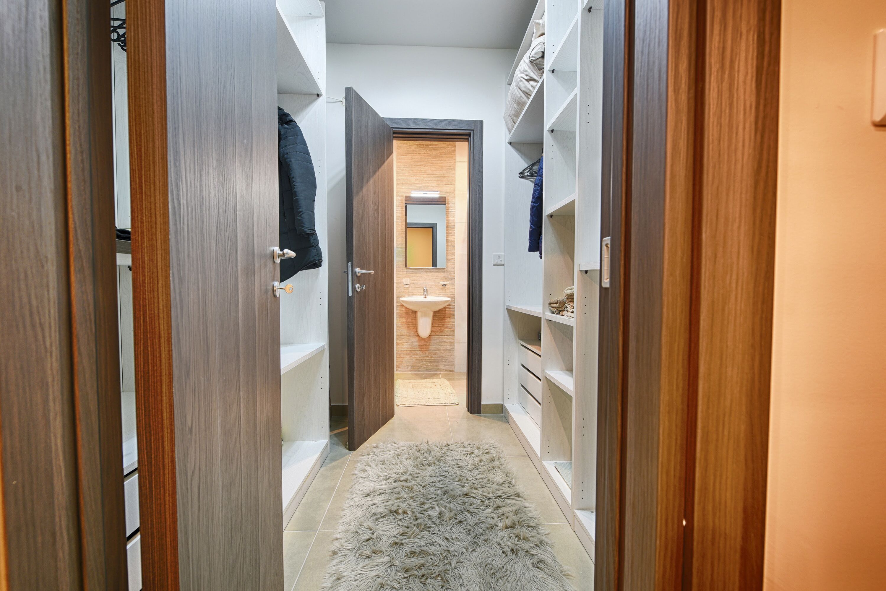 A narrow hallway with wooden doors and white shelving units, leading to a bathroom with a sink and mirror.