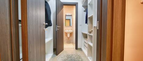 A narrow hallway with wooden doors and white shelving units, leading to a bathroom with a sink and mirror.