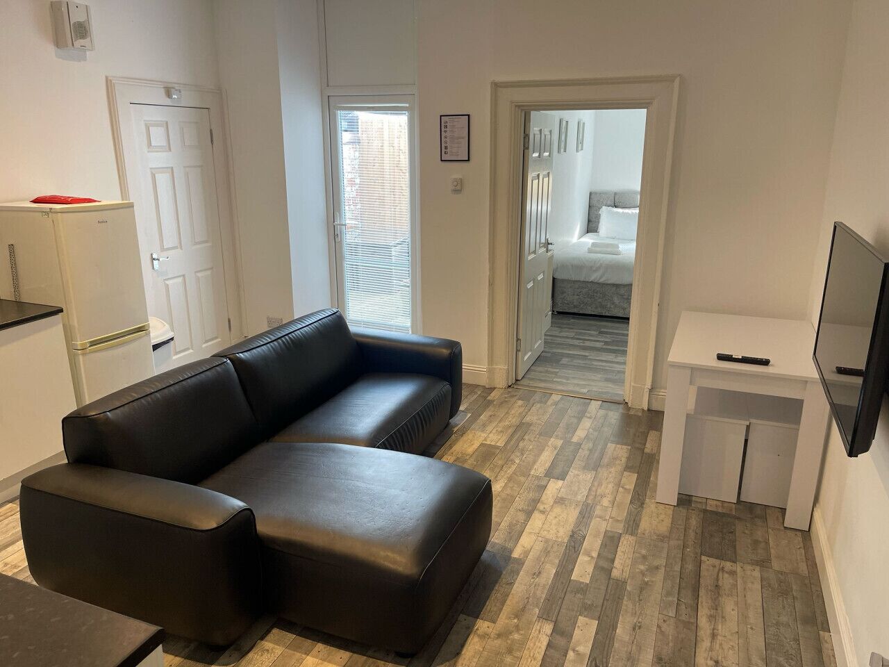Una sala moderna con un sofá de cuero negro, un mueble bajo para televisor blanco y suelo de parquet de madera.