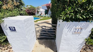 A gated entrance with white walls, numbered 19 and 217, leading to a pool area with lounge chairs and a clear view of a building with a red-tiled roof. - Villa Cristina, air conditioning, private pool, free WiFi (Binibeca)