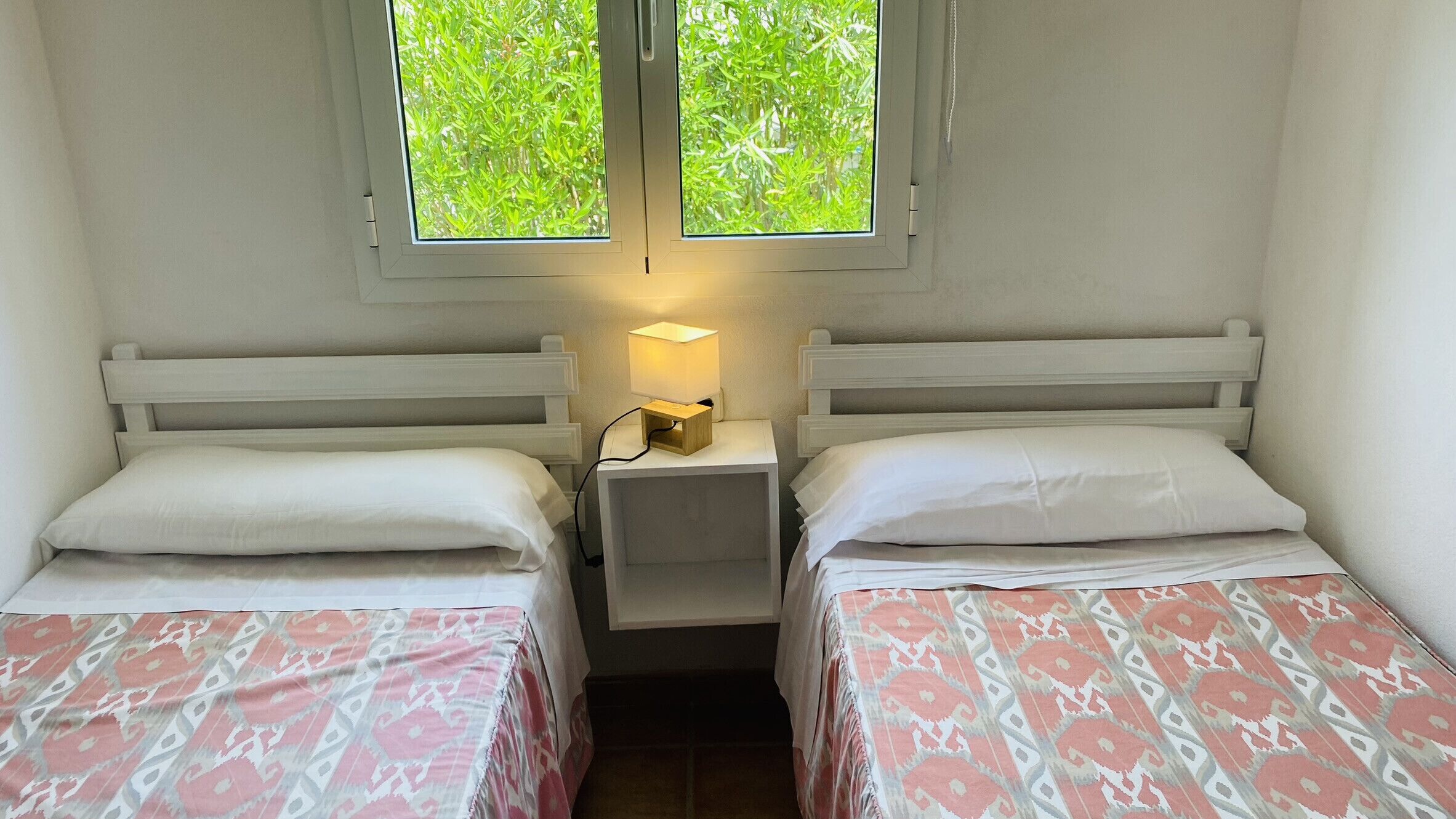 Ein kleines, sauberes Zimmer mit zwei Einzelbetten, einem Nachttisch mit Lampe und einem Fenster mit Blick auf Grünflächen.