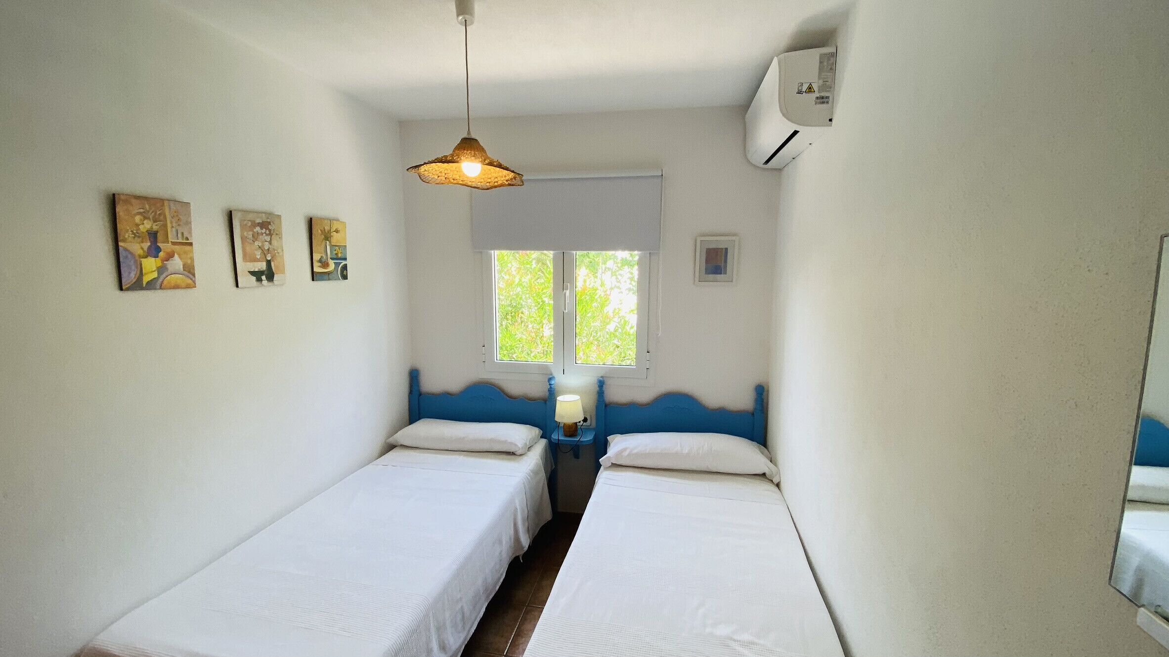 Quarto com duas camas individuais, janela com vista para área verde, luminária pendente e ar-condicionado fixado na parede.