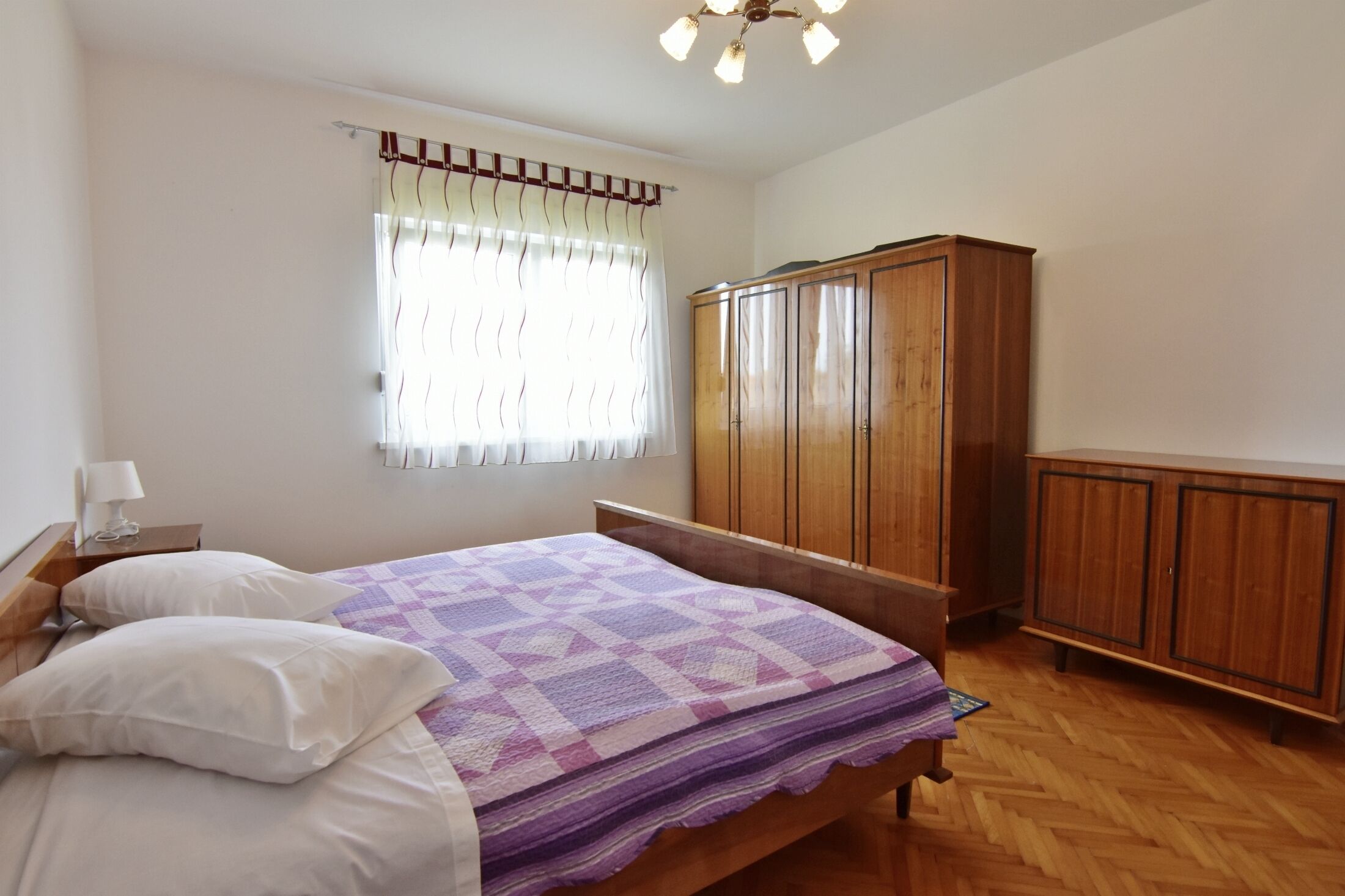 Ein Schlafzimmer mit einem hölzernen Bett, einem Nachttisch mit Lampe, einem Kleiderschrank und einem Fenster mit Vorhängen.
