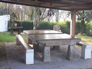 Une terrasse extérieure avec une table en pierre et des bancs, entourée de verdure et d’arbres.