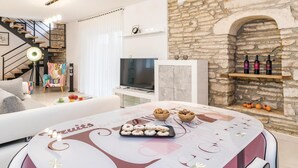 A modern living room with a stone fireplace, a dining table with a dessert display, and a TV in the background. - Amazing home in Sajini (Sajini)