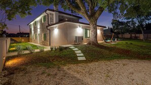 A two-story house with a stone facade, a visible air conditioning unit, and a well-maintained lawn with a stone pathway leading to the entrance. - Amazing home in Sajini (Sajini)