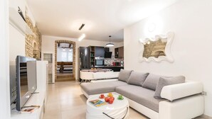A modern living room with a white sofa, a coffee table with books and fruit, a TV, and a kitchen in the background. - Amazing home in Sajini (Sajini)