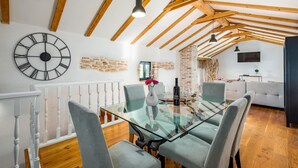 Un coin repas avec une table en verre, des chaises à revêtement gris, une grande horloge murale et des poutres en bois apparentes.