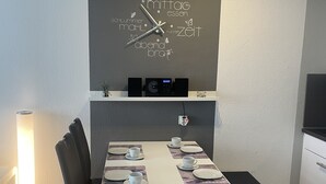 Et moderne spiseområde med et hvidt bord dækket til fire personer, sorte stole og et vægur.