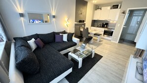 En moderne stue med en mørkegrå hjørnesofa, et glasbord og et køkken i baggrunden.