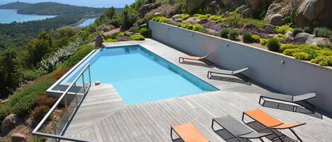 Piscina moderna al aire libre con cubierta de madera, rodeada de exuberante vegetación y con vistas a un paisaje costero pintoresco.