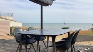 Een overdekt terras met tafel en stoelen, met uitzicht op een strand.