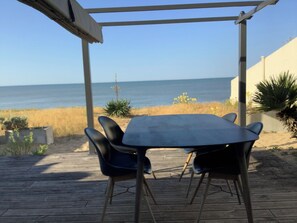 Eine Holzterrasse mit Tisch und Stühlen, die auf einen Strand hinausführt.