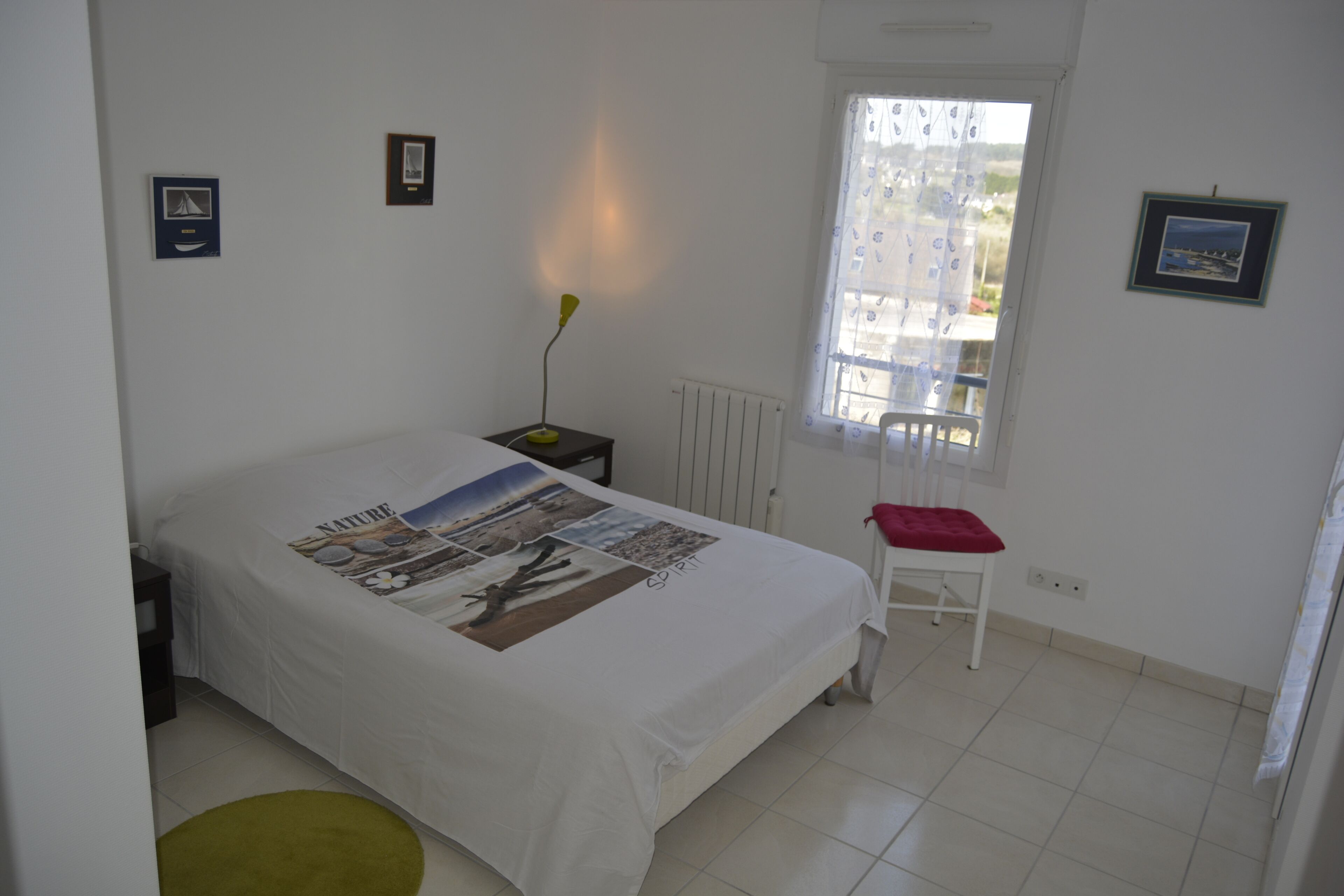 A bedroom with a bed, a nightstand, a chair, a window, and wall-mounted pictures.