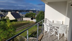 Un balcon donnant sur une ville côtière, avec des meubles d’extérieur blancs et une rambarde en métal.
