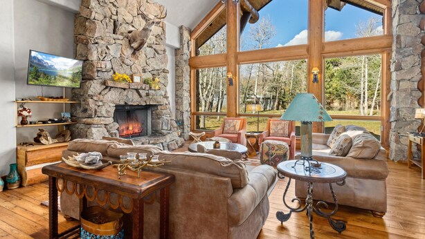 Un salon chaleureux avec une cheminée en pierre, des sièges confortables et de grandes fenêtres donnant sur l’extérieur.