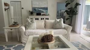 Un salon avec un canapé blanc, des coussins décoratifs, une table basse ornée d’un centre de table en forme de pomme de pin, et un tableau accroché au-dessus du canapé.