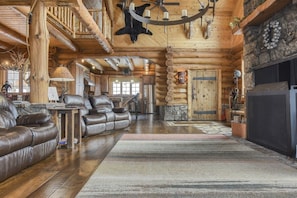 A log cabin interior with a fireplace, leather sofas, a chandelier, and antlers on the ceiling.