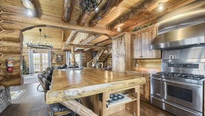 A log cabin interior with a large wooden dining table, a built-in stove, and a staircase leading to an upper level. - Stone Lodge-Panoramic Views, Lots of Space! (Lead)