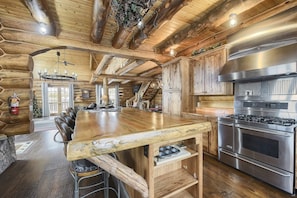 A log cabin interior with a large wooden dining table, a built-in stove, and a staircase leading to an upper level.
