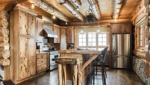 A rustic cabin-style kitchen with wooden cabinetry, a central island with stools, a stainless steel refrigerator, and a stone fireplace. - Stone Lodge-Panoramic Views, Lots of Space! (Lead)