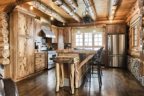 A rustic cabin-style kitchen with wooden cabinetry, a central island with stools, a stainless steel refrigerator, and a stone fireplace.