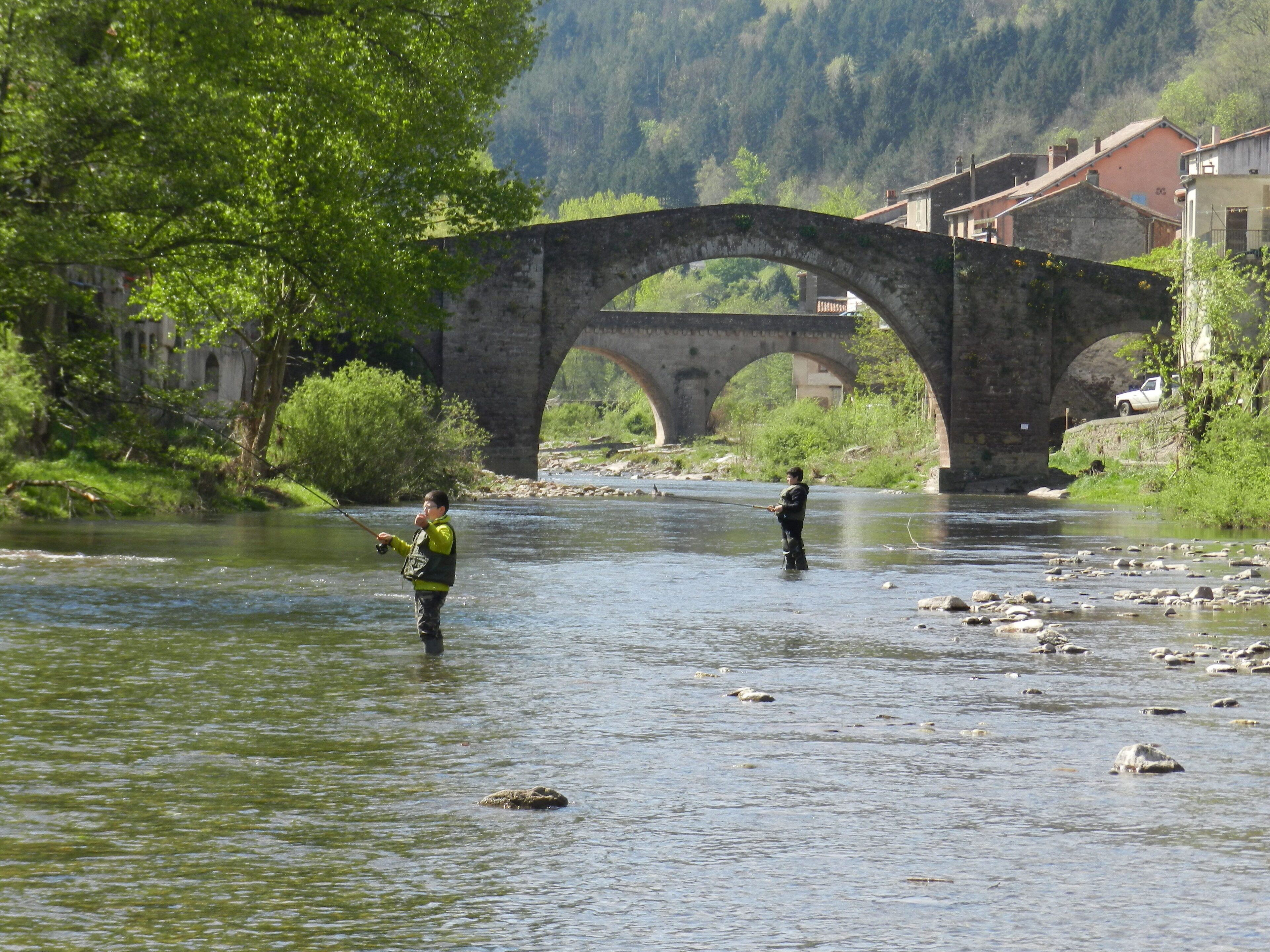 Une rivière où deux personnes pêchent, un pont de pierre et des bâtiments en arrière-plan.