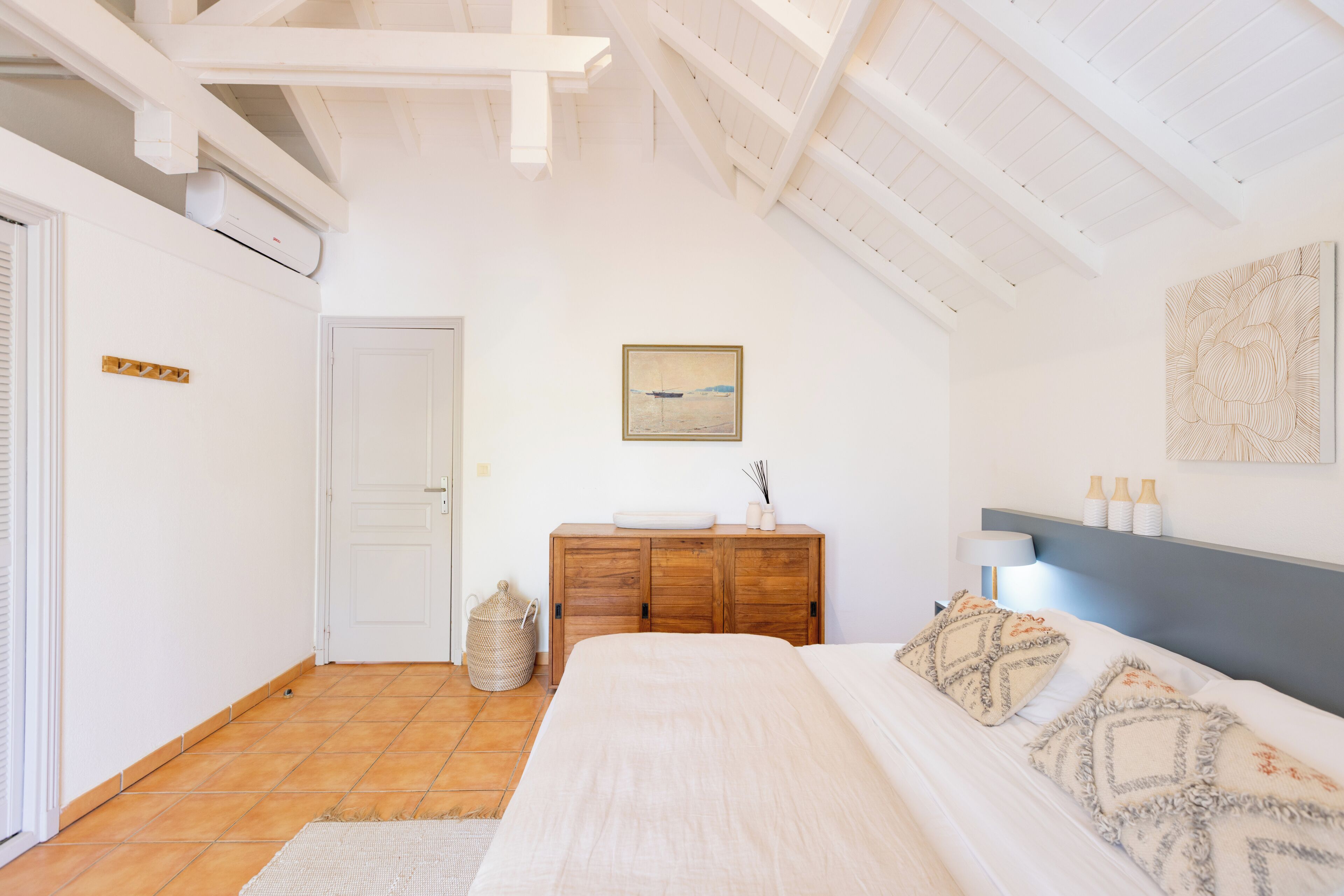 A bedroom with a bed, a wooden dresser, a painting, and a ceiling with exposed beams.