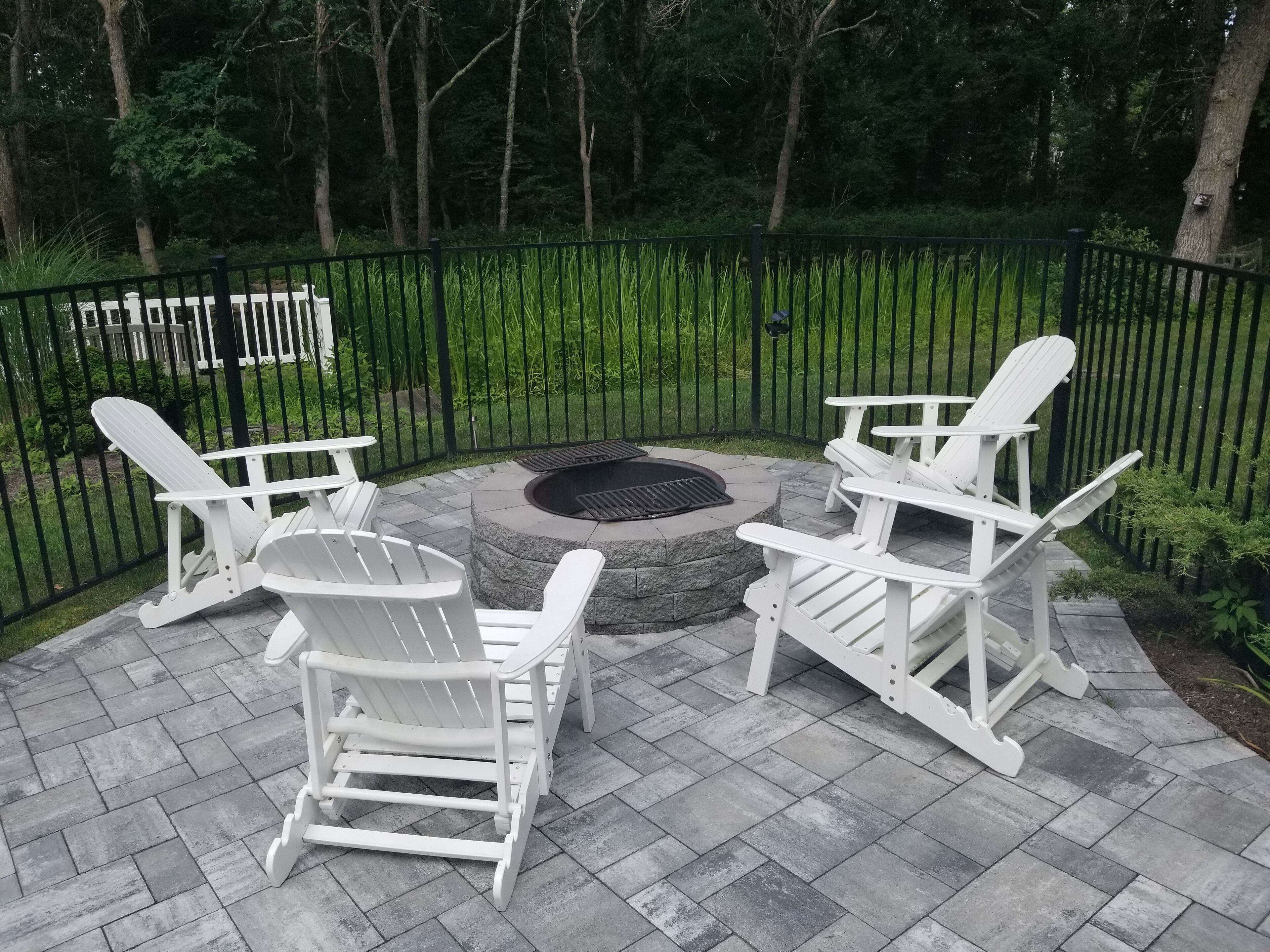 A patio area with Adirondack chairs, a circular stone fire pit, and a black metal fence.