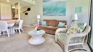 Ein Wohnzimmer mit einem braunen Sofa, weißen Korbstühlen, einem Glastisch mit rosa Blumen und einem großen Wandbild einer Welle.