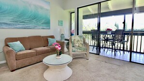 Ein Wohnzimmer mit beigefarbenem Teppich, einer braunen Couch, einem weißen Couchtisch und einem Korbstuhl. Eine große Glastür führt zu einem Außenbereich mit Tisch und Stühlen.