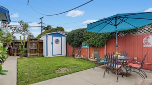 Un jardin avec un petit abri de jardin, une table de patio avec des chaises et un parasol bleu.