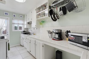 Een keuken met witte kastjes, een fornuis, een koffiezetapparaat en hangende pannen.