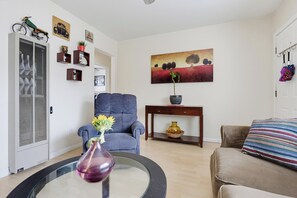 Un salon avec un fauteuil bleu, une table ronde ornée d’un vase, un canapé et un tableau accroché au mur.