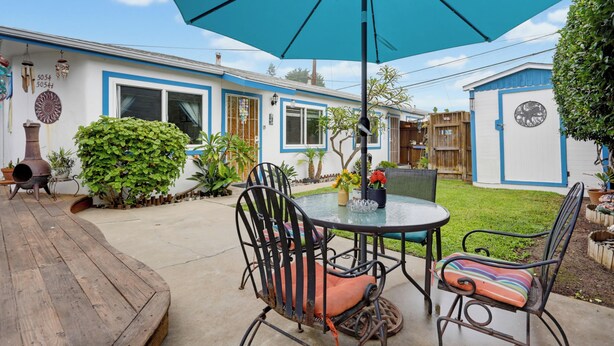 Une terrasse avec une table et des chaises, un parasol bleu, et une maison aux bordures bleues et à la porte blanche.