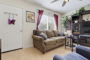 Een woonkamer met een beige bank, een tafeltje, een tv-meubel en een raam met gordijnen.