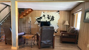 A wooden interior with a sofa, a coffee table, and a chair. There is a staircase with a wooden railing and a lamp on the floor.