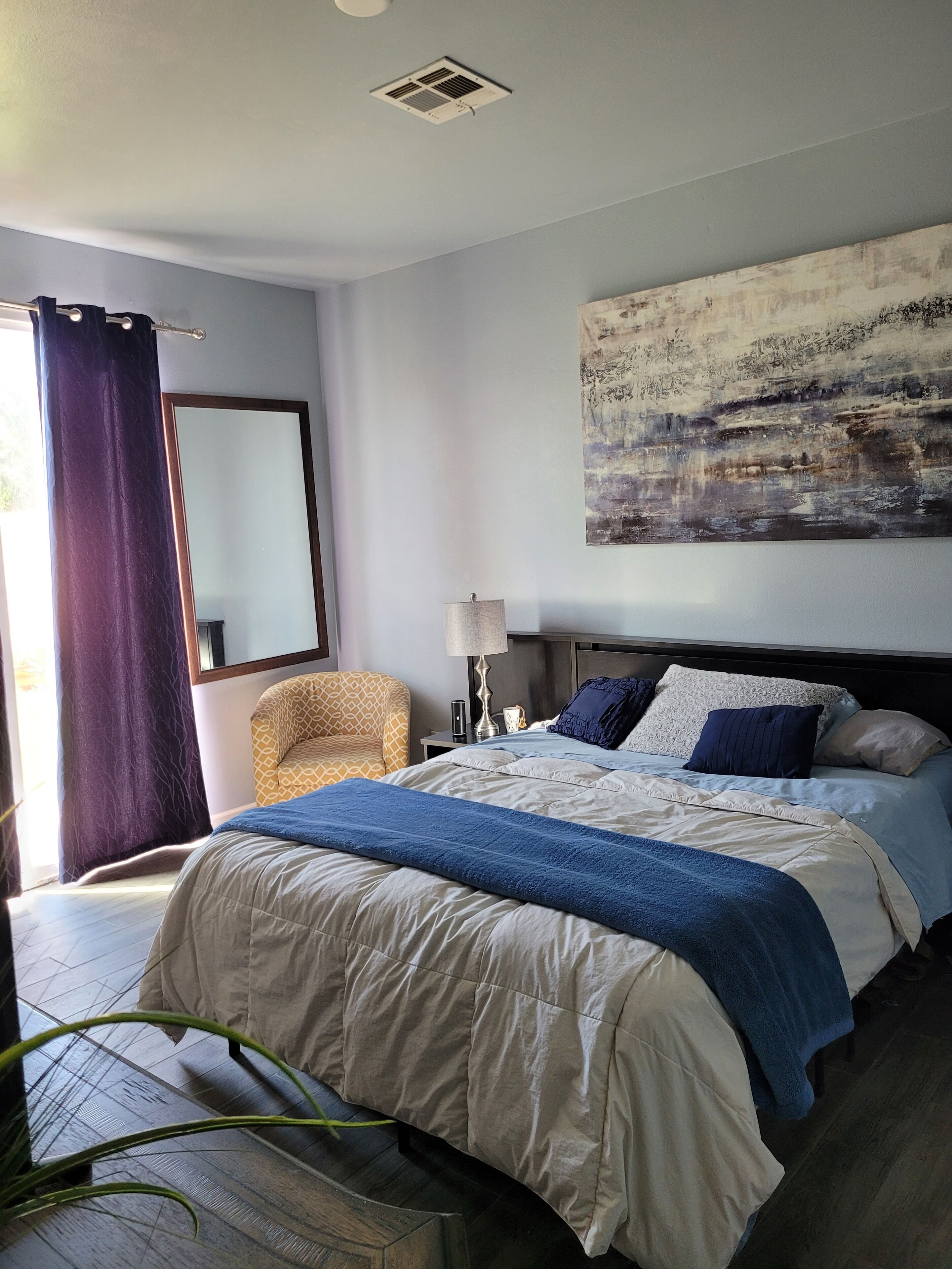 A bedroom with a large bed, a chair, a nightstand, a lamp, and a painting on the wall.