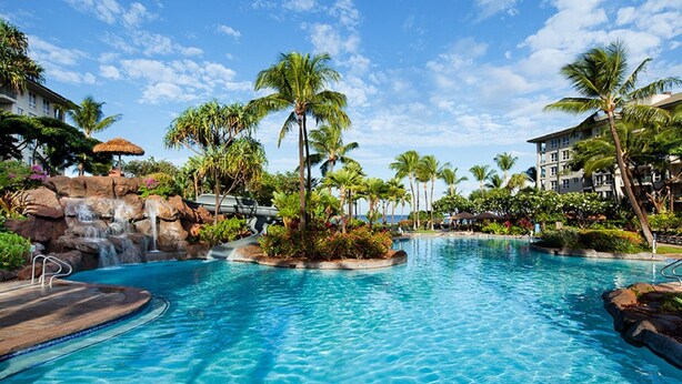 Une grande piscine extérieure agrémentée d’une cascade, entourée de palmiers et de plantes tropicales, avec des bâtiments en arrière-plan.