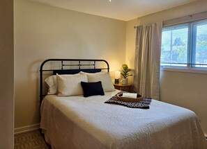 Een netjes opgemaakt bed met een hoofdeinde, een nachtkastje met een lamp en een raam met gordijnen.