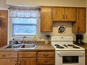 A kitchen with wooden cabinets, a double sink, a stove, and a window with blinds. - Yellowstone Vacation Condo (Miles City)