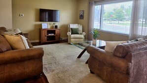 A living room with two sofas, a glass coffee table, a television on the wall, and a window with blinds.