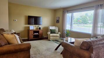 A living room with two sofas, a glass coffee table, a television on the wall, and a window with blinds.