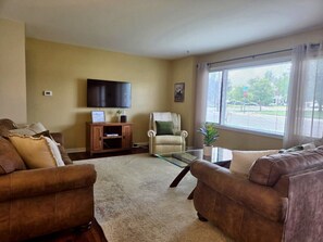 A living room with two sofas, a glass coffee table, a television on the wall, and a window with blinds. - Yellowstone Vacation Condo (Miles City)