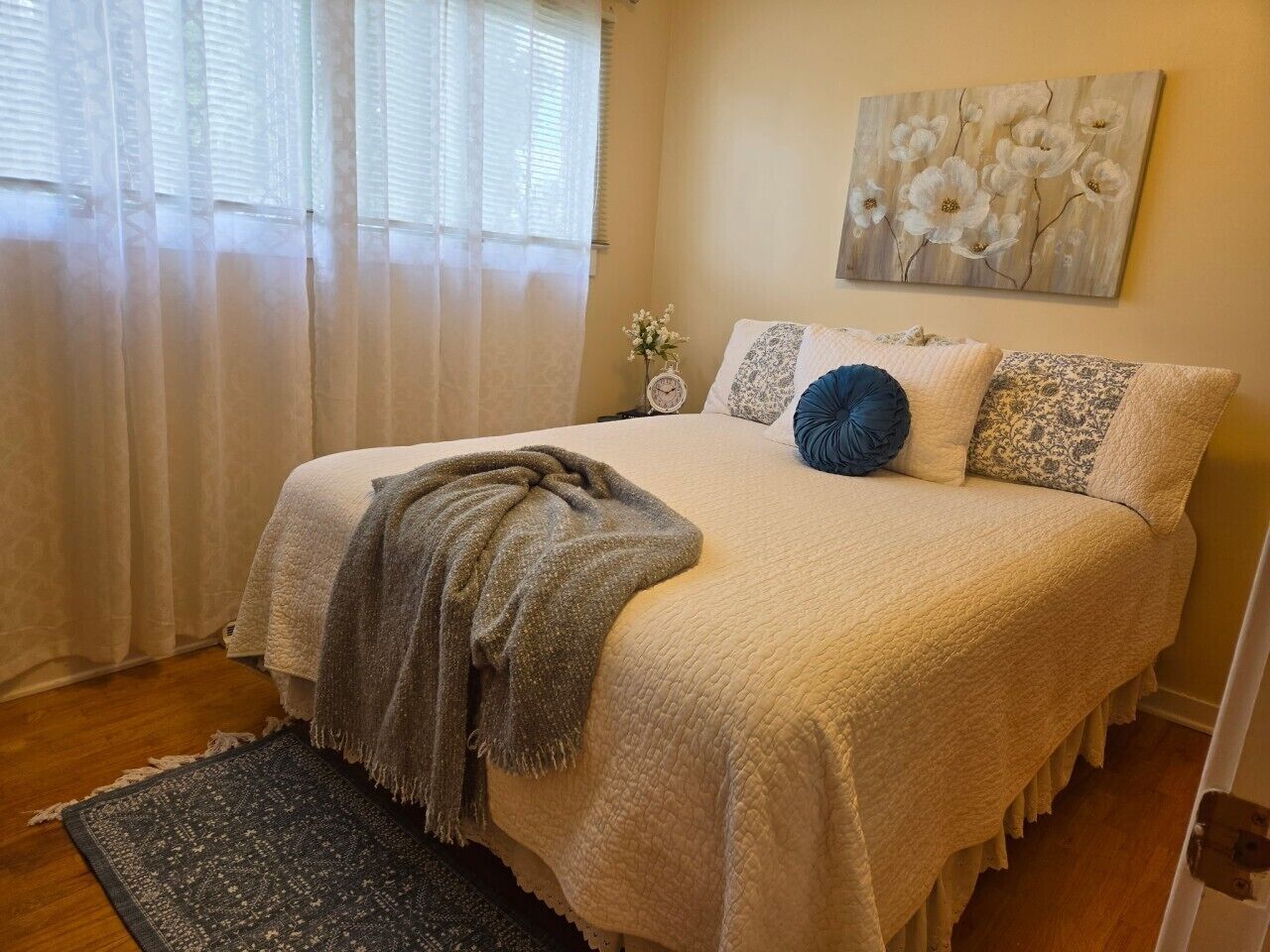 A bedroom with a bed, a bedside table, a nightstand, a window with curtains, and a wall-mounted painting.