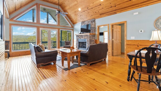 A spacious living room with a stone fireplace, wooden flooring, and large windows offering a view of the outdoors.